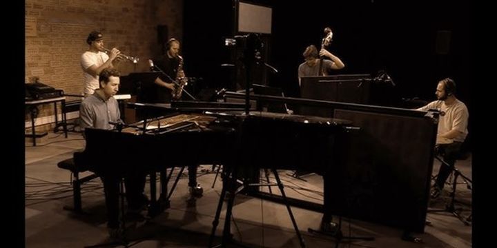 Quintett sitzt zusammen und musiziert, Flügel im Vordergrund, dahinter Trompete- und Saxophonspieler