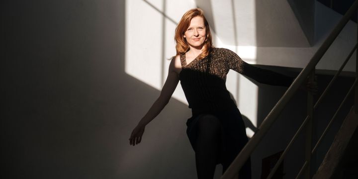 rothaarige Frau auf einer Treppe, hält sich mit dem linken Arm am Treppengeländer fest, rechtes Bein steht angewinkelt auf dem Treppengeländern, beide Arme sind ausgestreckt