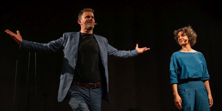 Mann, Tom van Mieghem, vorln links stehend, Frau, Antje Horn im Hintergrund rechts stehend, während einer Aufführung