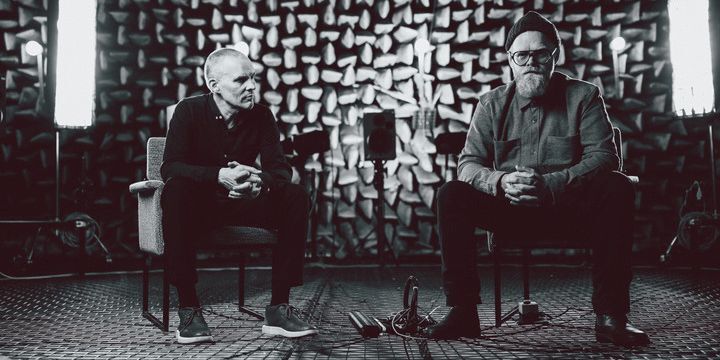 Schwarz-Weiß, zwei Männer sitzen im Raum, einer schaut in die Kamera, der andere sieht zu dem anderen Mann hinüber.